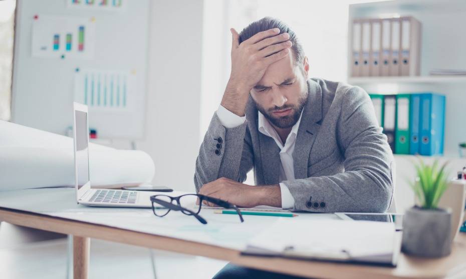 A person at an office desk resting their head on one hand beside an open laptop and paperwork